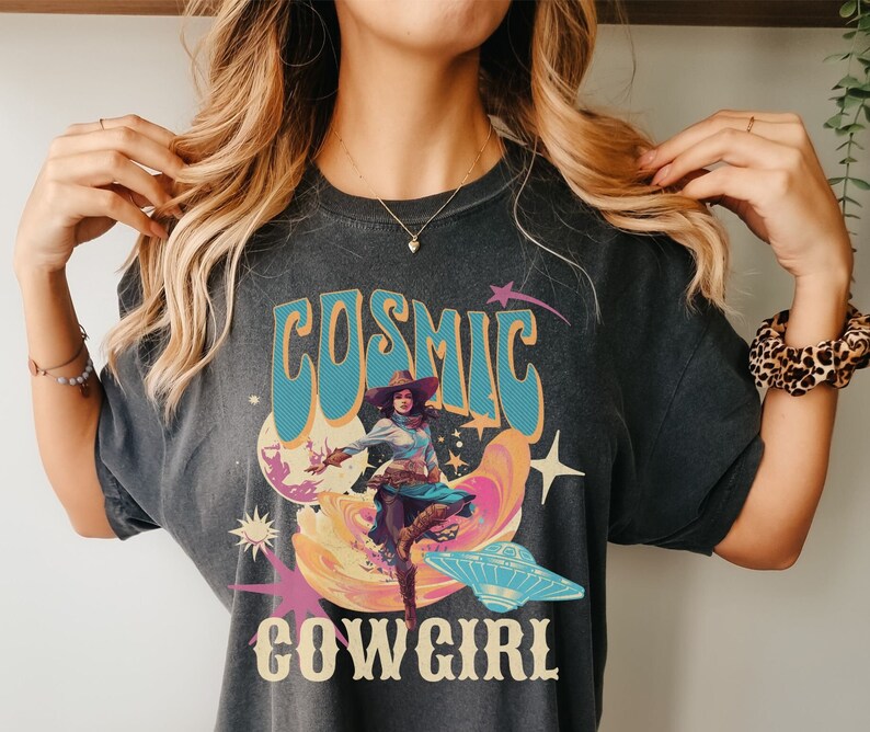 May include: A dark grey t-shirt with a retro graphic design featuring a woman in a cowboy hat riding a cosmic wave. The text "COSMIC COWGIRL" is printed on the shirt.