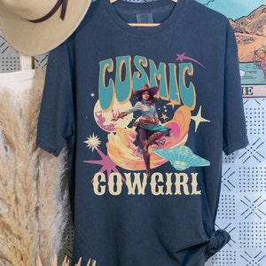May include: A dark blue t-shirt with a retro graphic design featuring a cowgirl riding a UFO with the text "Cosmic Cowgirl".