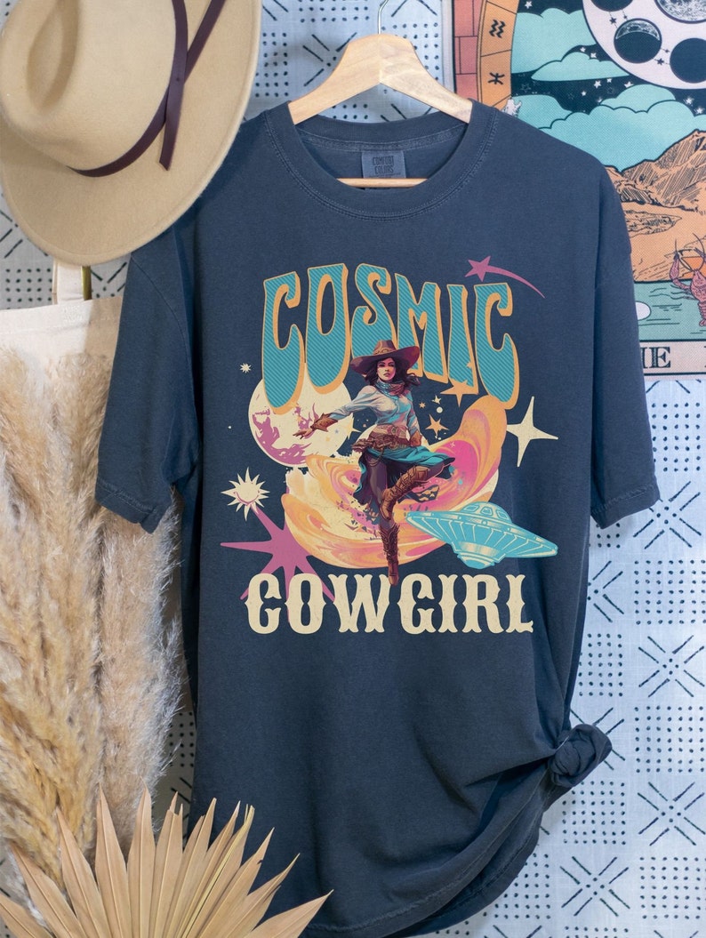 May include: A dark blue t-shirt with a retro graphic design featuring a cowgirl riding a UFO with the text "Cosmic Cowgirl".