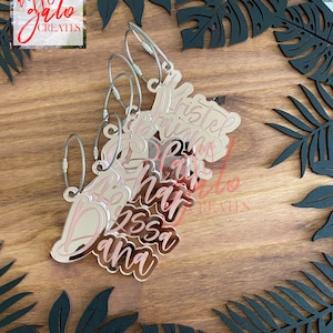 May include: Five silver keychains with pink lettering. The keychains are on a wooden surface with black leaf-shaped cutouts. The keychains are personalized with the names "Krista", "Brianna", "Blair", "Jessa", and "Ana".