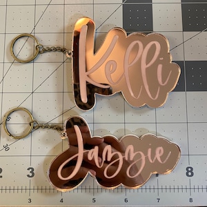May include: Two rose gold mirror acrylic keychains with the names "Kelli" and "Jazzie" written in white lettering.