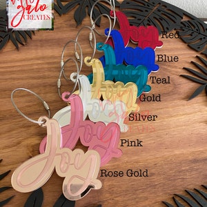 May include: A set of eight "Joy" keychains in various colors, including rose gold, pink, silver, gold, teal, blue, and red. Each keychain features a mirrored finish and a silver cable chain.