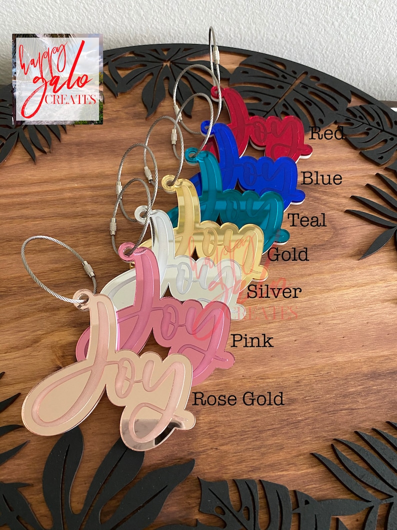 May include: A set of eight "Joy" keychains in various colors, including rose gold, pink, silver, gold, teal, blue, and red. Each keychain features a mirrored finish and a silver cable chain.