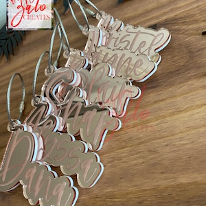 May include: A set of eight rose gold acrylic keychains with names written in white lettering. The keychains are attached to silver metal rings.
