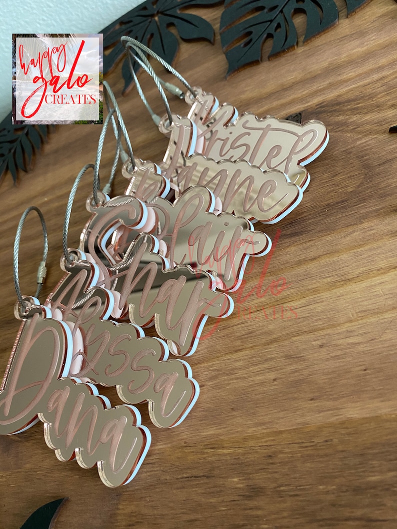 May include: A set of eight rose gold acrylic keychains with names written in white lettering. The keychains are attached to silver metal rings.
