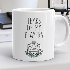 May include: White ceramic coffee mug with black text that reads "Tears of my players" and a black and white illustration of a twenty-sided die with a crown on top and green floral accents.