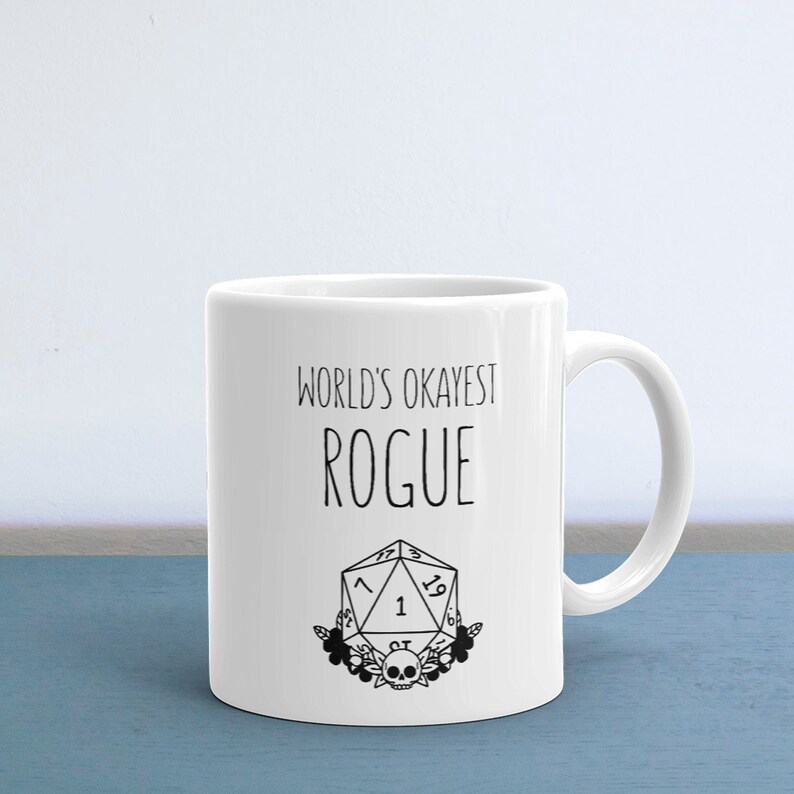 May include: White ceramic mug with a black design that says "World's Okayest Rogue" and a black and white illustration of a twenty-sided die with flowers and a skull.