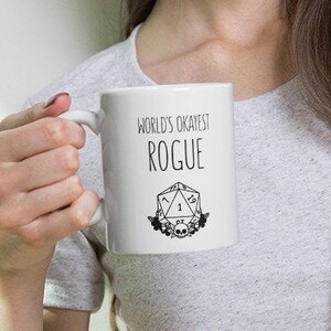 May include: White ceramic mug with a black design that says "World's Okayest Rogue" and a black and white illustration of a twenty-sided die with a skull and crossbones.