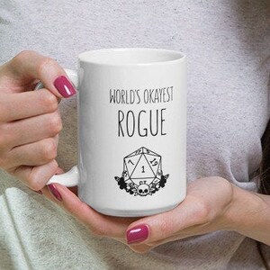 May include: White ceramic mug with a black design that says "World's Okayest Rogue" and a black and white illustration of a 20-sided die with a skull and flowers.