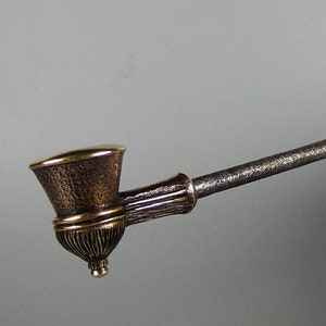 May include: A long, thin, metal pipe with a decorative, ornate bowl. The bowl is made of brass and has a gold finish. The pipe is held in a hand with red nail polish.