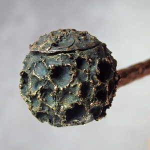 Puede incluir: Una pequeña escultura redonda de bronce con una superficie texturizada. La escultura está unida a un palo de madera.