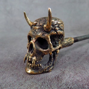 Puede incluir: Una pipa de cráneo de bronce detallada con cuernos, el cráneo es texturizado y tiene una sonrisa de dientes. La pipa está unida a un tallo largo de metal oscuro.