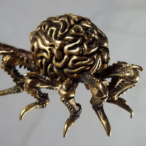 Pode incluir: Uma escultura de metal dourado de uma criatura com forma de cérebro e seis patas. A criatura é detalhada com texturas intrincadas e parece estar rastejando.
