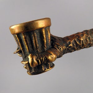 Può includere: Una pipa di bronzo a forma di mano che tiene una ciotola. La ciotola è testurizzata e ha un bordo dorato. La mano è dettagliata con artigli e rughe.