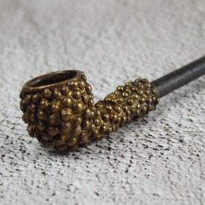 May include: A close-up of a vintage tobacco pipe. The pipe features a brass-colored bowl and stem, covered in small, textured bumps. The stem is a dark, matte color. The pipe is set against a neutral background.