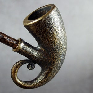 May include: A brass pipe with a hammered finish and a decorative, curled handle. The pipe is made of metal and has a rustic, antique look.