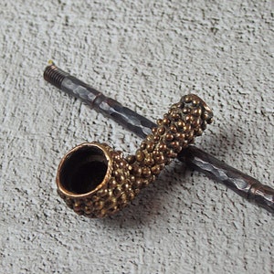 May include: A handcrafted tobacco pipe with a textured, bronze-colored bowl and a dark, hammered metal stem. The bowl is covered in small, rounded protrusions. The stem is resting on a textured, light gray surface.