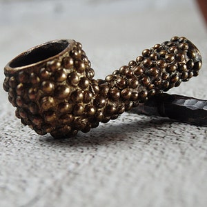 May include: A vintage-style tobacco pipe with a textured, brass-colored finish. The bowl and stem are covered in small, rounded bumps, giving it a unique, tactile appearance. The pipe is designed with a curved stem and a dark, open bowl.