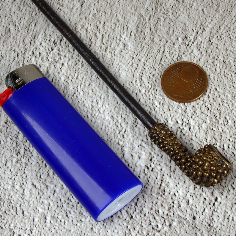 May include: A blue lighter, a small bronze pipe with a textured bowl and stem, and a bronze coin are arranged on a textured surface. The pipe has a dark, slender stem and a detailed, rounded bowl.
