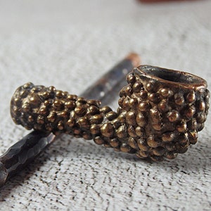 May include: A close-up of a bronze-colored tobacco pipe with a textured, bumpy surface. The pipe's bowl and stem are visible, showcasing a detailed, handcrafted design. The pipe is resting on a light-colored surface.