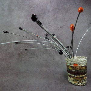 May include: A ceramic vase with a textured, multi-colored surface, featuring a pattern of brown, orange, and white. The vase holds a sculpture of thin metal wires with black and orange beads.