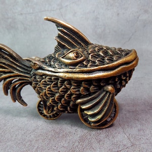 Könnte beinhalten: Eine bronzene, stilisierte Fischskulptur mit filigranen Details, darunter Schuppen und Flossen. Der Fisch ist auf Rädern und hat einen skurrilen, fast cartoonartigen Ausdruck.