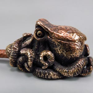Puede incluir: Escultura de bronce de un pulpo con detalles intrincados. La escultura presenta una cabeza y tentáculos detallados, con un acabado texturizado. El pulpo está montado sobre una varilla de color cobre, creando una pieza decorativa única.