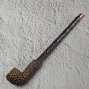 May include: A handcrafted pipe with a dark, textured stem and a brass-colored bowl. The bowl has a textured, dotted pattern. The end of the stem has a copper-colored tip. The pipe is designed for smoking.