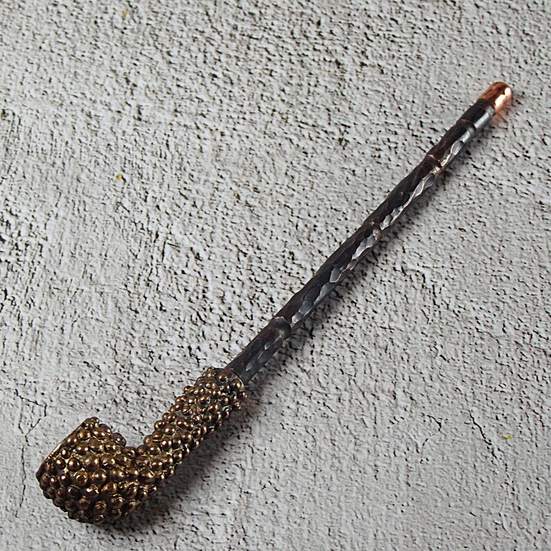 May include: A handcrafted pipe with a dark, textured stem and a brass-colored bowl. The bowl has a textured, dotted pattern. The end of the stem has a copper-colored tip. The pipe is designed for smoking.