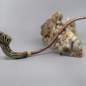 May include: A decorative pipe with a brass bowl and a copper stem. The bowl is shaped like a stylized flower with multiple layers. The stem is textured and has a gold accent near the bowl.