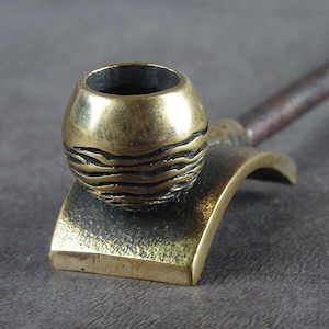 May include: A brass pipe with a round bowl and a curved stem. The bowl has a textured design of wavy lines.