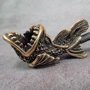 May include: A bronze-colored bottle stopper featuring a detailed fish design with an open mouth. The stopper has a textured finish and is designed to fit into a bottle. The fish's scales and fins are intricately carved.