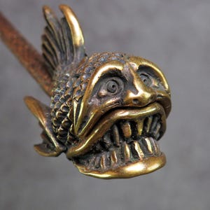 Könnte beinhalten: Ein messingfarbenes, fischförmiges Deko-Objekt mit detailliertem, offenem Gesicht und scharfen Zähnen. Der Fisch hat Flossen und Schuppen und ist an einem braunen Stab befestigt. Das Objekt hat ein Vintage- oder Antik-Aussehen.