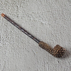 May include: A handcrafted pipe with a dark, textured stem and a bulbous bowl. The bowl and the area where the stem meets the bowl are adorned with a textured, golden-brown pattern. The end of the stem has a copper-colored tip.