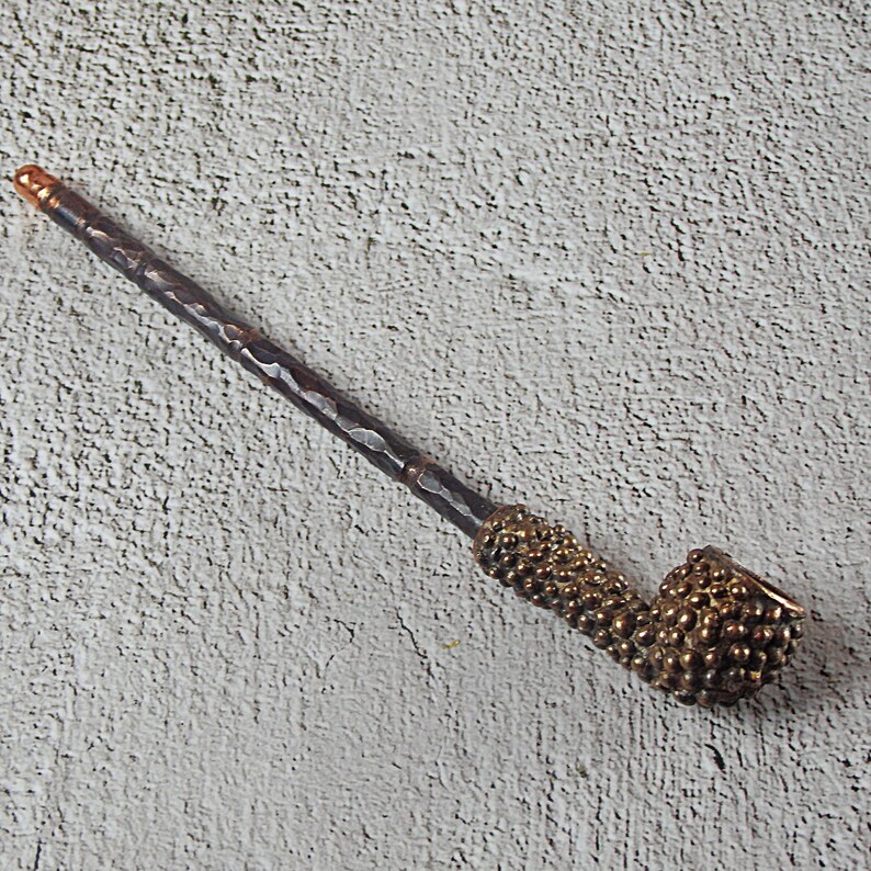 May include: A handcrafted pipe with a dark, textured stem and a bulbous bowl. The bowl and the area where the stem meets the bowl are adorned with a textured, golden-brown pattern. The end of the stem has a copper-colored tip.