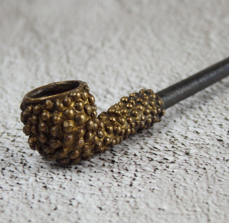 May include: A close-up of a vintage tobacco pipe. The pipe features a brass-colored bowl and stem, covered in small, textured bumps. The stem is a dark, matte color. The pipe is set against a neutral background.