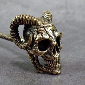 May include: A detailed bronze-colored skull with curved horns and a long, textured handle. The skull has visible teeth and eye sockets, and the horns curve upwards. The overall design suggests a gothic or fantasy theme.