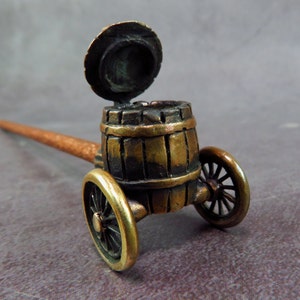 May include: A small, antique-looking, bronze-colored pipe with a wooden stem and a barrel-shaped bowl on wheels. The bowl is open and the lid is propped up.