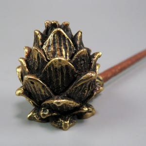 Puede incluir: Una pipa de latón con un cuenco en forma de flor y un tallo marrón. El cuenco tiene un diseño detallado y texturizado con un acabado dorado. El tallo tiene una forma cilíndrica y delgada.