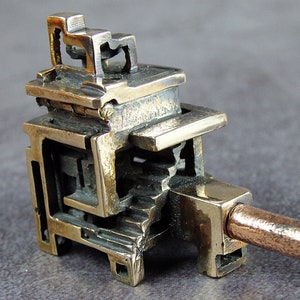May include: A bronze-coloured, abstract sculpture of a miniature structure with geometric shapes and a copper rod extending from the side. The sculpture has a detailed, textured surface and a unique, artistic design.