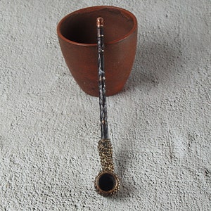 May include: A handcrafted, ornate pipe with a dark bowl and a textured, hammered metal stem. The stem has copper accents and a decorative gold-colored band near the bowl. The pipe rests near a brown ceramic mug, suggesting a smoking accessory.
