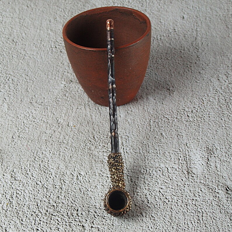 May include: A handcrafted, ornate pipe with a dark bowl and a textured, hammered metal stem. The stem has copper accents and a decorative gold-colored band near the bowl. The pipe rests near a brown ceramic mug, suggesting a smoking accessory.