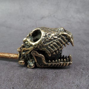 Könnte beinhalten: Eine detaillierte, bronzefarbene Skulptur eines Dinosaurierschädels mit offenem Maul, das scharfe Zähne zeigt. Der Schädel ist an einem strukturierten, goldbraunen Griff befestigt. Das Stück ist wahrscheinlich ein Dekorationsartikel oder ein einzigartiges Werkzeug.