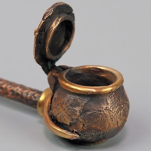 Op de afbeelding: Een klein, bronzen, scharnierend doosje met een open deksel. Het doosje heeft een getextureerd oppervlak en een gouden rand. Het doosje is bevestigd aan een lange, getextureerde, bronzen handgreep.
