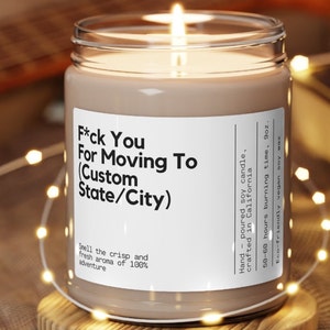 May include: A lit soy wax candle in a glass jar with a white label. The label reads "F*ck You For Moving To (Custom State/City)". The candle is 9oz and has a 50-60 hour burn time. The candle is surrounded by string lights.