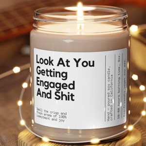 May include: A brown glass candle jar with a white label that reads "Look At You Getting Engaged And Shit". The candle is lit and the jar is surrounded by string lights.
