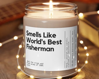 Op de afbeelding: Een bruine sojawas-kaars met een wit label waarop staat "Smells Like World's Best Fisherman". De kaars staat op een houten onderzetter en is omringd door witte lichtslingers.