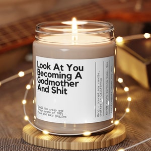May include: A brown soy candle with a white label that reads "Look At You Becoming A Godmother And Shit". The label also says "Smell the crisp and fresh aroma of 100% love and baby giggles". The candle is on a wooden coaster.