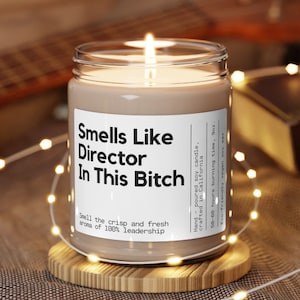 May include: A lit candle in a clear glass jar with a white label that reads "Smells Like Director In This Bitch." The candle sits on a wooden coaster, surrounded by string lights. The text below the title says "Smell the crisp and fresh aroma of 100% leadership."
