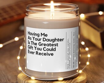 May include: A brown soy candle in a clear glass jar with a white label that reads "Having Me As Your Daughter Is The Greatest Gift You Could Ever Receive". The candle is sitting on a wooden coaster and surrounded by string lights.